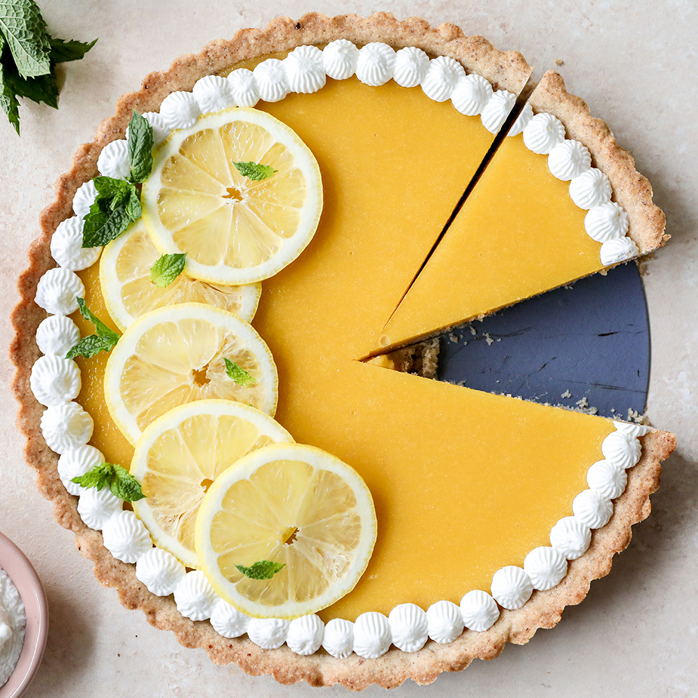 Lemon tart with a slice removed, topped with sliced lemons, mint leaves, and whipped cream border on a light background.
