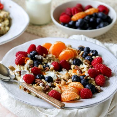 A bowl of yogurt topped with raspberries, blueberries, mandarin orange slices, and chopped nuts, served with a spoon, with more fruit in the background.