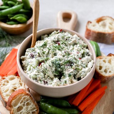 A bowl of creamy dip with herbs, surrounded by sliced baguette, carrot sticks, and snap peas on a wooden board.