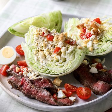 Plate with iceberg lettuce wedge salad, cherry tomatoes, blue cheese, crumbled bacon, hard-boiled egg, and slices of grilled steak.
