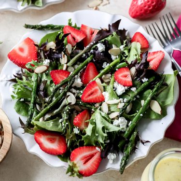 Fresh salad with mixed greens, sliced strawberries, asparagus, crumbled cheese, and almond slices on a white plate, accompanied by a whole strawberry and fork in the background.