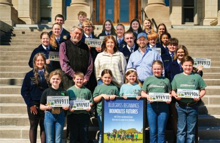 A diverse group of children and adults pose together on outdoor steps, holding signs and smiling, with a building entrance in the background.