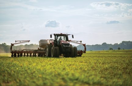 A tractor plows a green field under a blue sky with scattered clouds, preparing the land for planting on a rural farm.