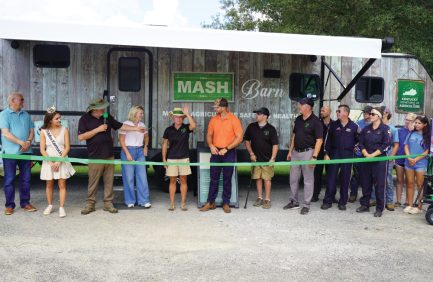 A group of people stands in front of a trailer, holding a green ribbon for a ribbon-cutting ceremony, with "MASH Barn" sign visible in the background.