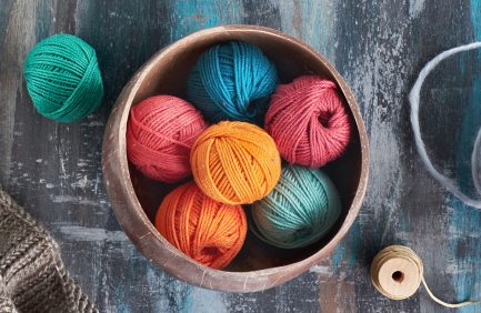 A wooden bowl filled with colorful yarn balls in orange, red, turquoise, and blue, placed on a textured, blue-painted surface.