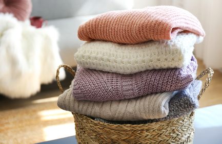 Four folded knit sweaters in pastel colors stacked in a wicker basket, with a cozy and sunlit background.