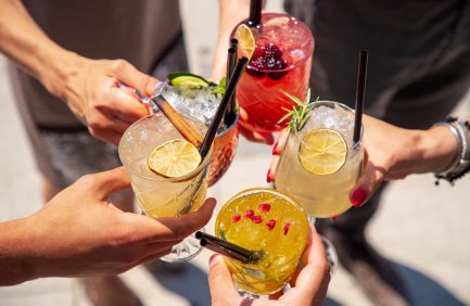 Five people raise colorful cocktails garnished with fruits and herbs, toasting together in bright sunlight.