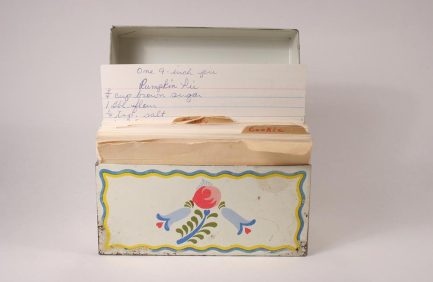 A vintage recipe box with a floral design and handwritten recipe cards inside, one card propped up and partially visible, against a plain background.