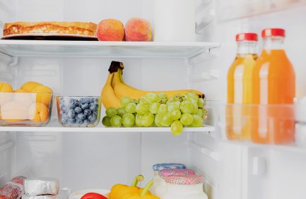 Open refrigerator with bananas, green grapes, blueberries, peaches, orange juice, a bell pepper, and yogurt on the shelves.