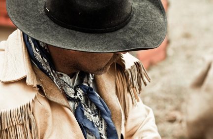 Man wearing a wide-brimmed black hat, tan fringed jacket, and blue patterned scarf, looking downward.