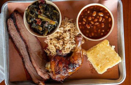 Barbecue plate with smoked brisket, pulled pork, chicken leg, baked beans, collard greens, and a slice of cornbread on a tray.