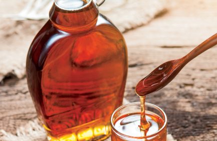 A glass bottle and jar filled with amber maple syrup, with syrup being poured from a spoon, on a wooden surface.