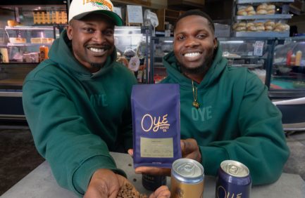 Two smiling men in green hoodies hold a bag of OYE coffee and display coffee beans and canned drinks on a counter.
