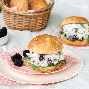 Sandwich with creamy chicken salad and blackberries on a bun, served on a pink plate with fresh blackberries, bread rolls in a basket in the background.