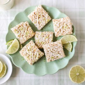 Six rice crispy treats are arranged on a green plate with lime wedges, on a light-colored tablecloth.