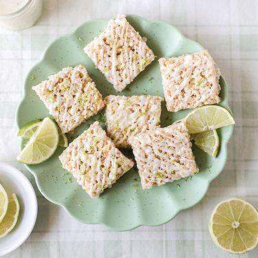 Six rice crispy treats are arranged on a green plate with lime wedges, on a light-colored tablecloth.