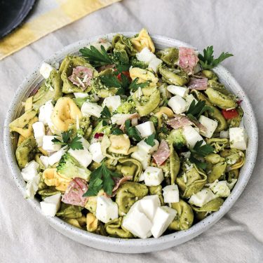 A bowl of tortellini pasta salad with diced cheese, ham, fresh herbs, and vegetables, served on a light cloth background.