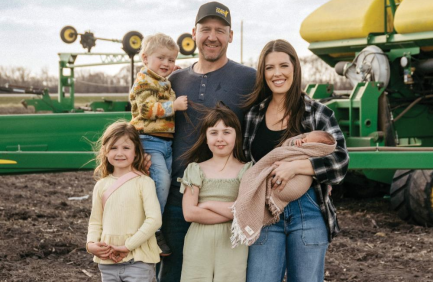 A family of five, including two adults and three young children, poses together outdoors near green farm equipment on a cloudy day.
