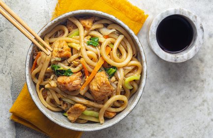 Bowl of stir-fried udon noodles with vegetables and chicken pieces, served with chopsticks and a cup of soy sauce on the side.