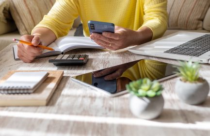 Person in yellow shirt using a smartphone, writing in a notebook, with a calculator, tablet, and a small potted plant on the table.