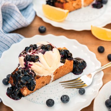 A slice of cake topped with whipped cream, blueberries, and banana slices on a white plate, with lemon wedges and extra blueberries nearby.