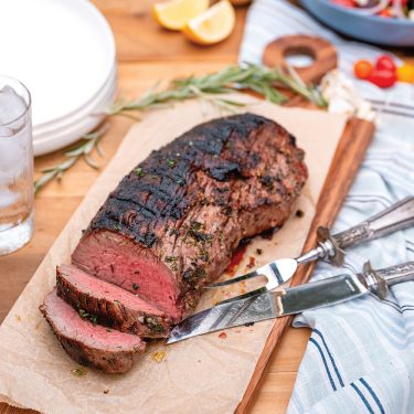 Sliced grilled steak resting on a wooden cutting board, garnished with herbs, with knives nearby and a plate of food and glass of water in the background.