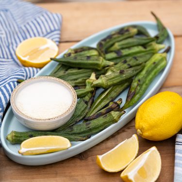 Grilled okra served on an oval plate with a bowl of dipping sauce, lemon wedges, and a whole lemon on a wooden table.