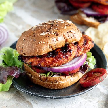 Grilled chicken sandwich with lettuce, tomato, and red onion on a whole wheat sesame bun, served on a black plate with greens and chips nearby.