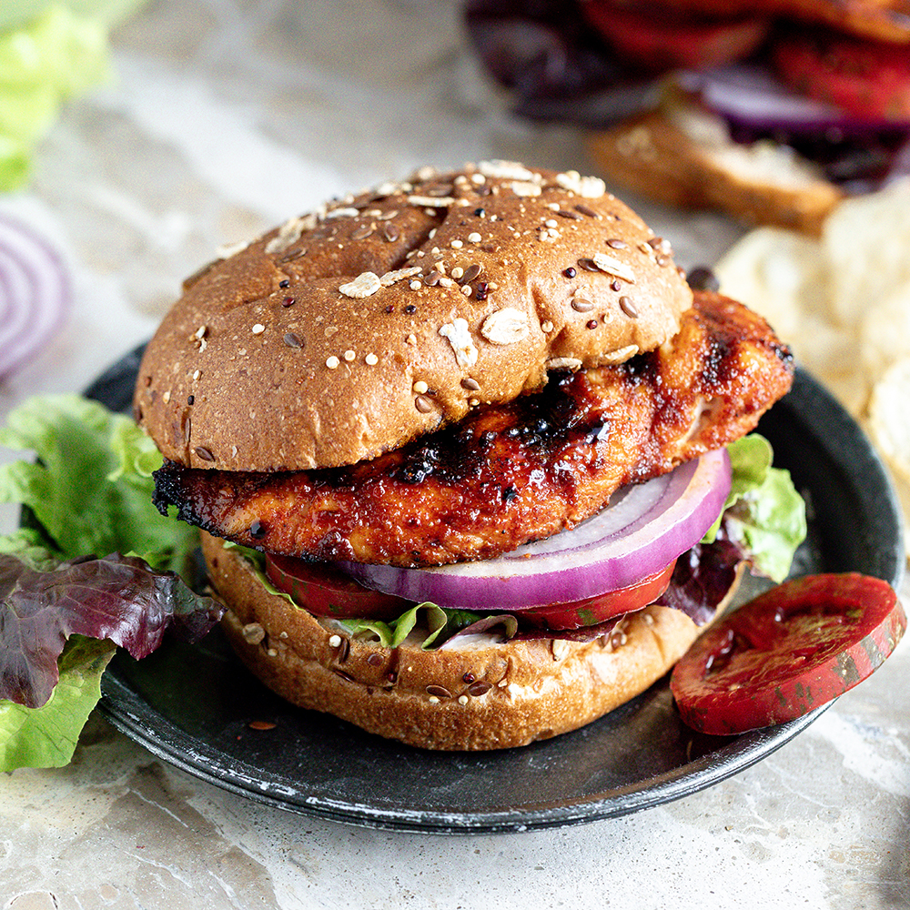 Grilled chicken sandwich with lettuce, tomato, and red onion on a whole wheat sesame bun, served on a black plate with greens and chips nearby.