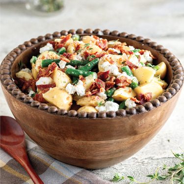 Wooden bowl filled with potato salad, green beans, bacon bits, and crumbled cheese, placed on a light-colored surface with a wooden spoon beside it.