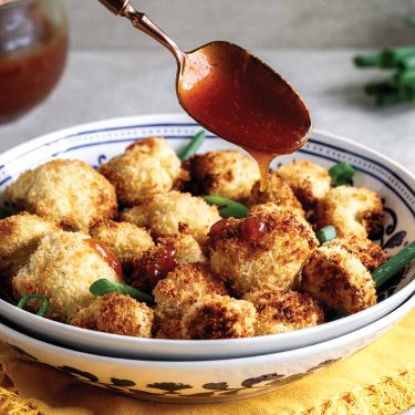 A bowl of crispy breaded cauliflower bites being drizzled with a reddish sauce, garnished with fresh herbs, served on a patterned plate with a yellow napkin underneath.