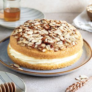 Round sponge cake filled with cream, topped with sliced almonds, served on a blue plate. A honey dipper and tea are visible in the background.