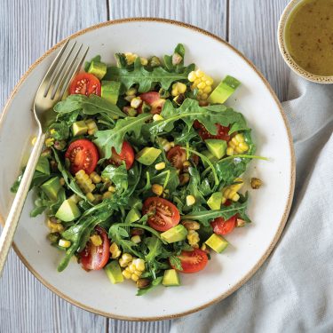 Fresh salad with arugula, cherry tomatoes, corn, avocado, and dressing served on a white plate with a fork, next to a cup of vinaigrette.
