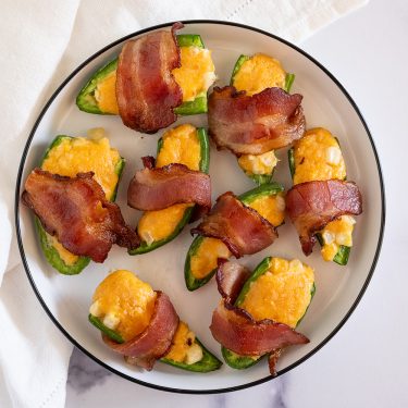 Plate of jalapeño poppers stuffed with cheese and wrapped in crispy bacon, arranged neatly on a round white plate.
