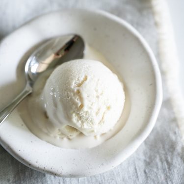 A scoop of vanilla ice cream in a white bowl with a silver spoon on a light-colored cloth.