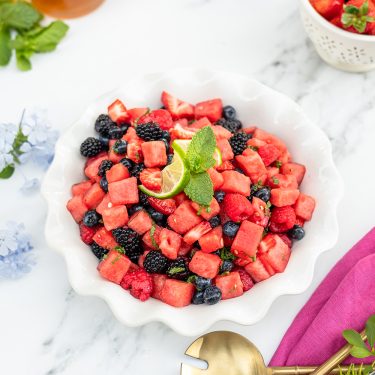 A white bowl filled with a colorful fruit salad containing watermelon cubes, blueberries, and blackberries, garnished with mint and lime slices on a marble surface.