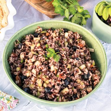 A bowl of mixed wild rice salad with chopped vegetables and herbs, garnished with fresh parsley, placed on a light-colored tablecloth.