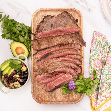 Sliced medium-rare steak on a wooden board with cilantro, avocado, salad, and a floral napkin on a white surface.