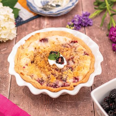 A berry crumble pie in a white dish, topped with cream and a mint leaf, surrounded by flowers and a bowl of blackberries on a wooden table.