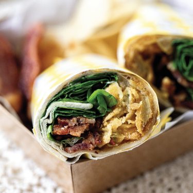 Close-up of a fresh wrap sandwich filled with greens, grilled chicken, and bacon, served in a cardboard container with chips in the background.