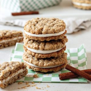 Stack of oatmeal cream pies with a creamy filling, placed on a green checkered paper, with cinnamon sticks and more pies in the background.