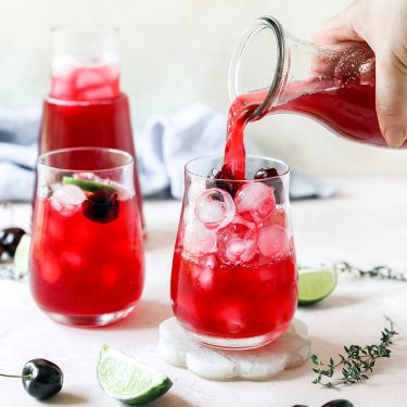 A hand pours a bright red drink with ice into a glass, garnished with cherries and lime, next to another glass and a carafe on a light surface.