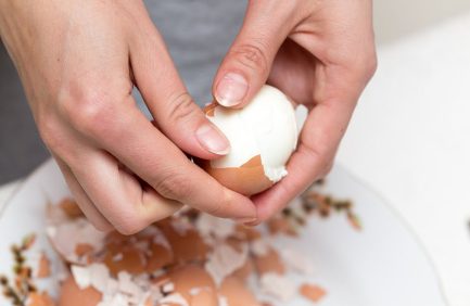 Hands peeling the shell off a hard-boiled egg over a plate with pieces of eggshell scattered around.