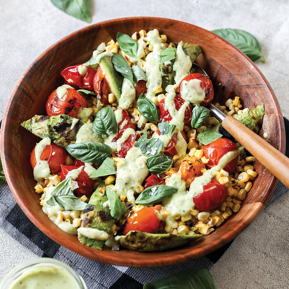 A wooden bowl filled with grilled vegetables, cherry tomatoes, corn, avocado slices, fresh basil leaves, and a creamy green dressing, with a fork on the side.