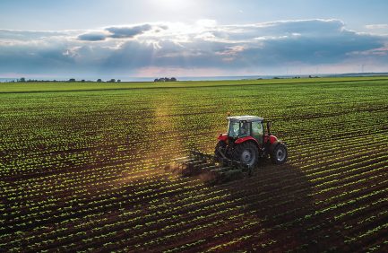 A red tractor plows a large, green crop field under a cloudy sky with sun rays shining through, creating a dynamic farming landscape.