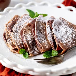 Chocolate crepes folded on a white plate, dusted with powdered sugar, garnished with fresh mint leaves, and served with a fork.