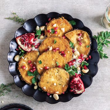 Fried eggplant slices on a black plate, garnished with pomegranate seeds, fresh herbs, chopped nuts, and pomegranate wedges.
