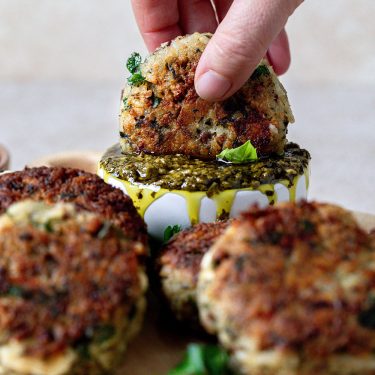 A hand dipping a crispy vegetable patty into green pesto sauce, with more patties and fresh herbs nearby.