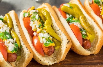 Chicago-style hot dogs topped with mustard, onions, relish, tomato slices, pickles, sport peppers, and a poppy seed bun on a wooden surface.