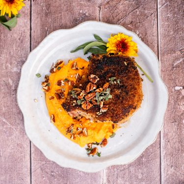 Breaded pork chop with mashed sweet potatoes, pecans, and herbs, served on a white plate garnished with a yellow flower and green leaves.